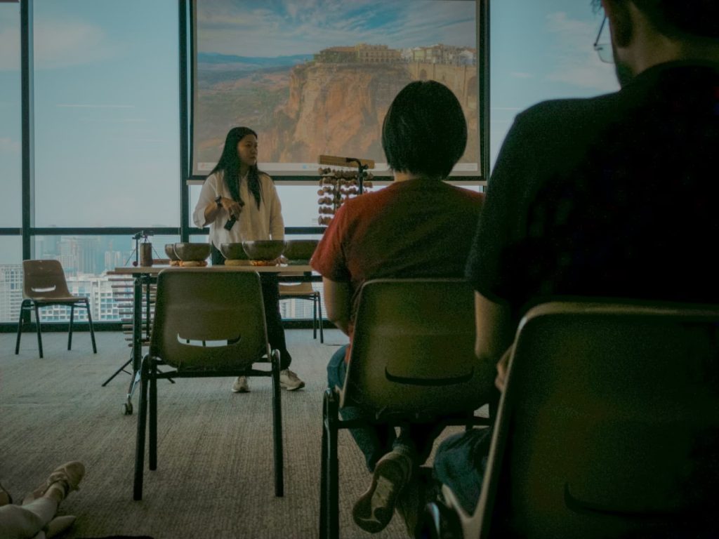 A facilitator standing at the front of a room holding a mallet during a sound bath session, viewed from behind seated participants in a university space in Singapore.