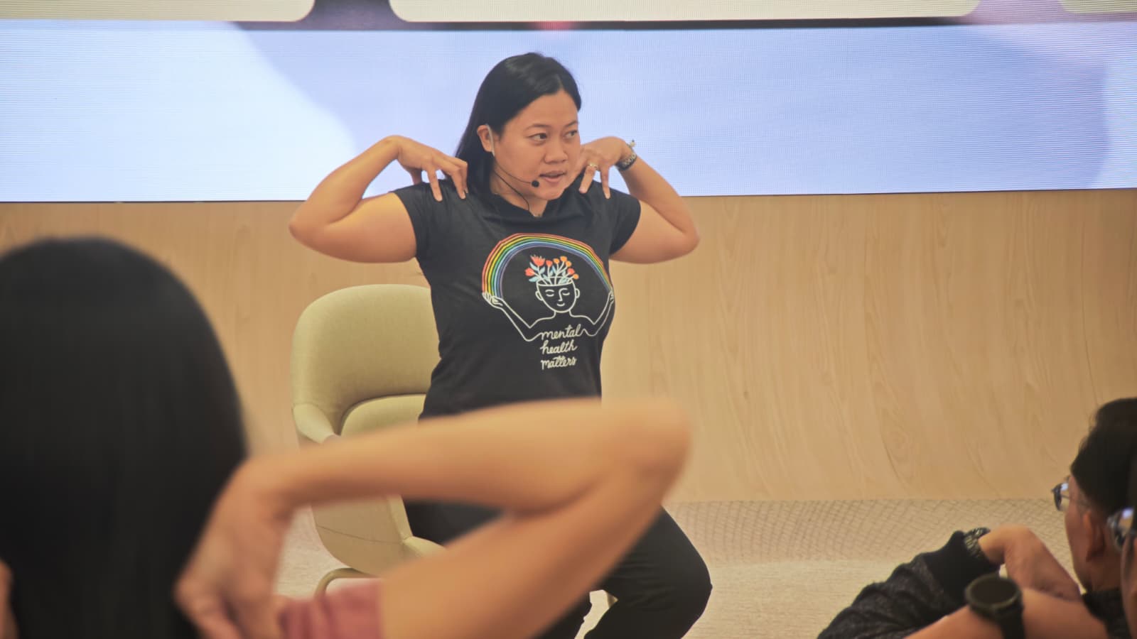 Wellness speaker guiding a corporate audience through gentle shoulder rolls and somatic movement exercises prior to a sound bath